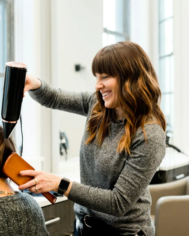 A smiling hair stylist blow-dries a client's hair in a bright salon.