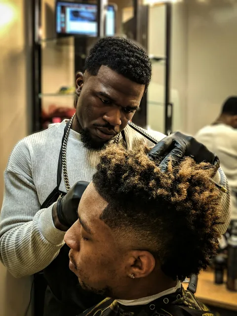 A barber in black gloves shapes a client's curly fade in a busy barbershop.