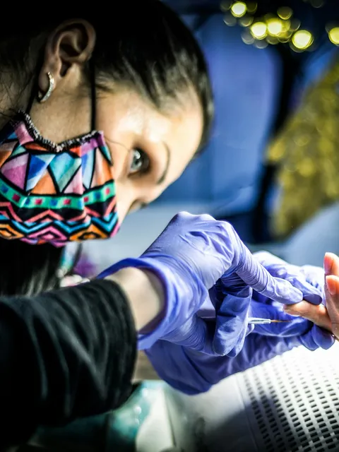 A nail technician in a mask and gloves works closely on a client's nails.
