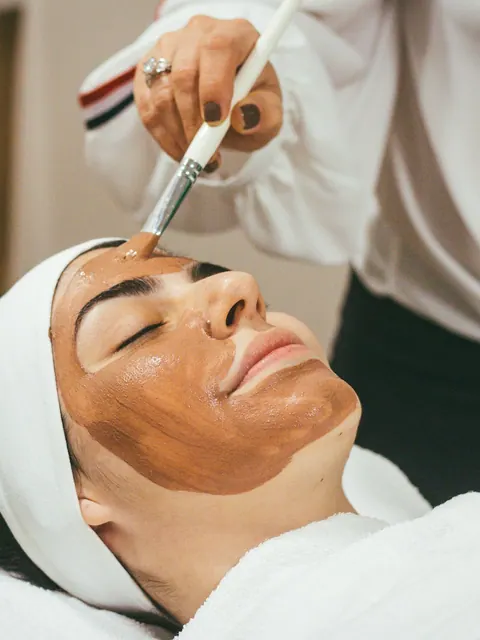 A therapist brushes a clay mask onto a relaxed client's face.